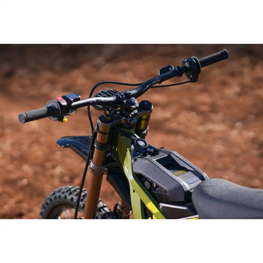 Close-up of a dirt bike handlebar area with black grips, wiring, a gray battery pack mounted on the frame, and a lime green frame, photographed outdoors against a blurred reddish-brown dirt background.