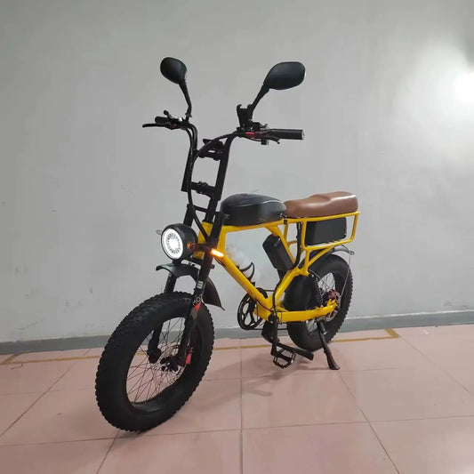 A yellow and black electric fat-tire bike with a brown saddle, black mirrors, and a headlight, standing on its kickstand in an indoor setting with a tiled floor and plain wall.