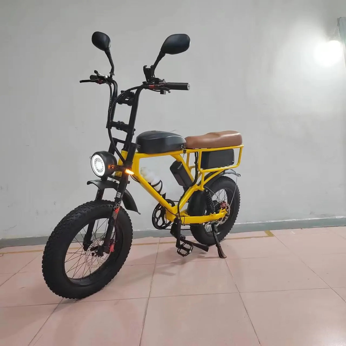 A yellow and black mini electric bicycle with fat tires, a brown saddle, and rear cargo rack, shown on a tiled floor against a plain wall.