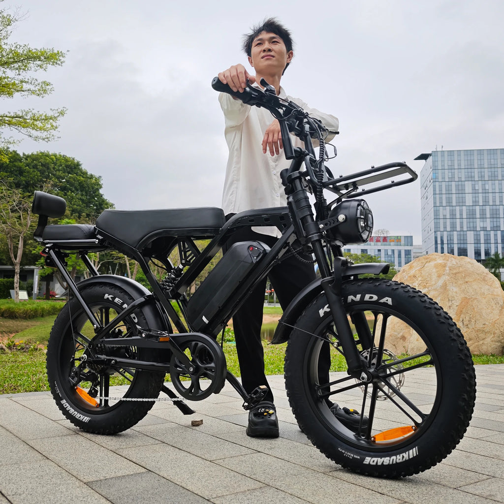 A man wearing a white shirt and black pants stands next to a black electric bicycle with fat tires, parked outdoors on a paved area with trees and a modern building in the background.