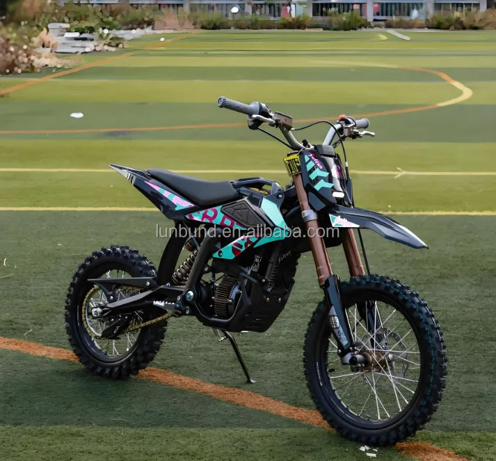 A small off-road dirt bike with black, gray, and turquoise graphics, parked on a green turf field with sports markings in the background.