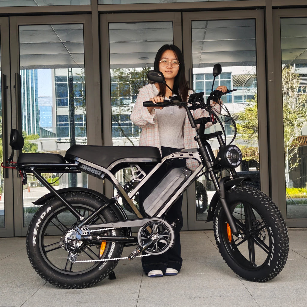 A woman stands behind a fat-tire electric bicycle in front of a glass building entrance. The bike has a black and silver frame with a rear cargo seat and thick knobby tires.