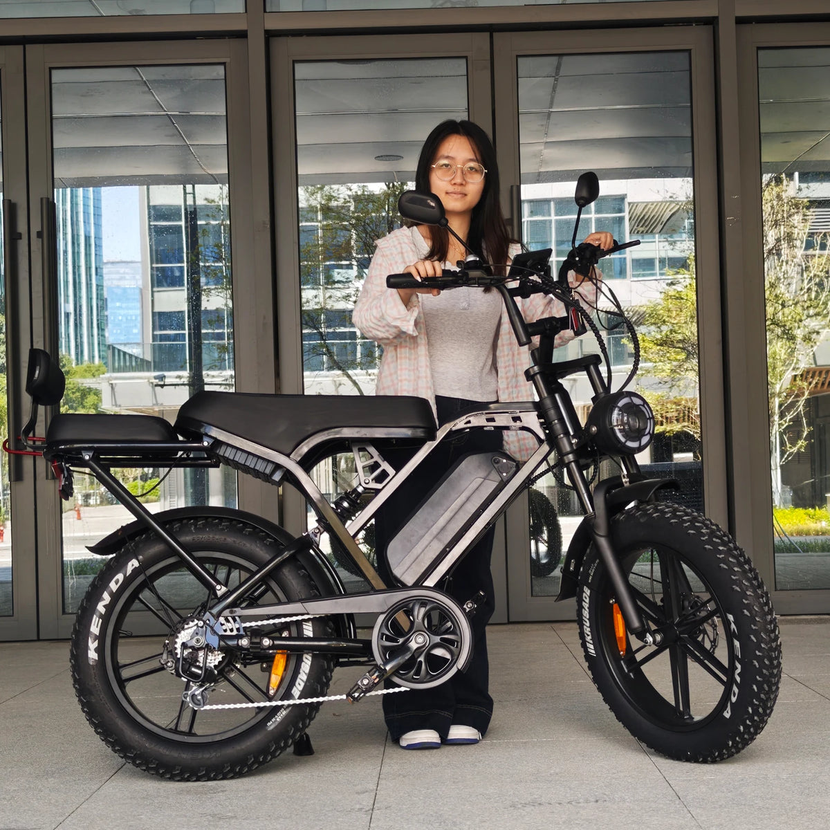A woman stands behind a fat-tire electric bicycle in front of a glass building entrance. The bike has a black and silver frame with a rear cargo seat and thick knobby tires.
