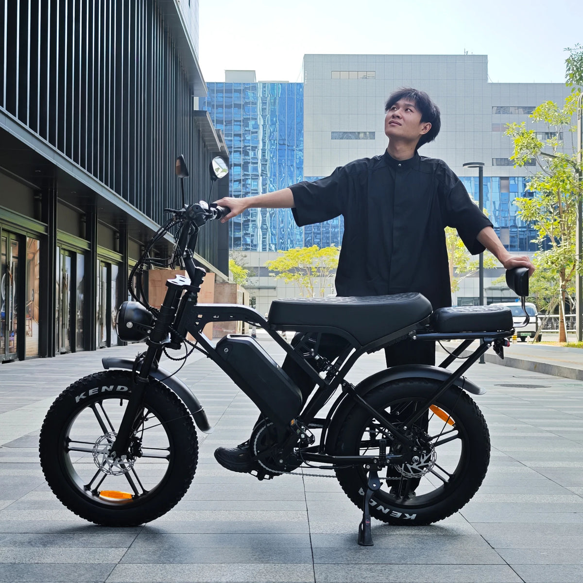A man in a black shirt stands beside a black electric bicycle on a paved outdoor walkway, with modern buildings and trees in the background.