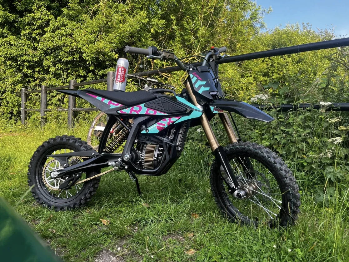 A black dirt bike with pink and teal graphics parked outdoors on grass with trees and a fence in the background. The bike has a silver canister bottle mounted on the handlebar.