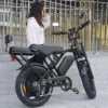 A woman wearing a white top and black skirt sits on a black electric scooter with a rear cargo bed, photographed outdoors against a blurred background of buildings and trees.