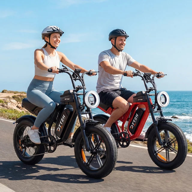 Two people riding electric bicycles on a coastal road with the ocean in the background. The bikes are branded OITN, with one bike in black and the other in red.