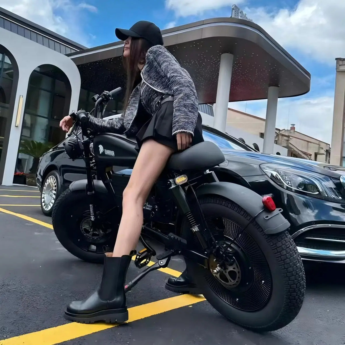 A woman wearing a black cap, gray marled jacket, black shorts, and black boots sits on a black electric motorcycle in a parking lot next to a black car.