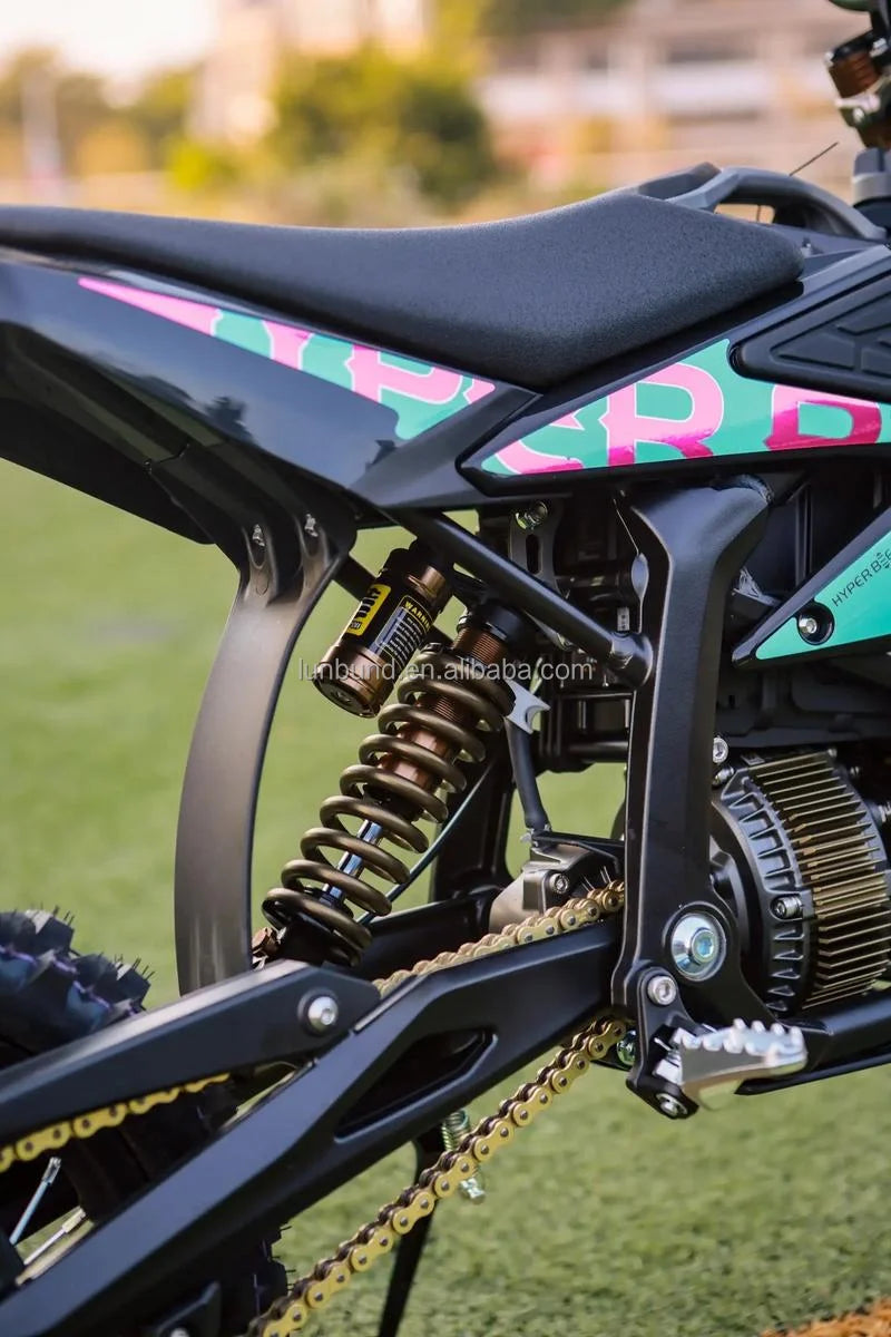 Close-up of a black dirt bike or motorcycle rear section with a gold chain, rear shock absorber, engine components, and a black seat. The side panel has pink and green graphic lettering, and the background shows blurred green grass and outdoor scenery.