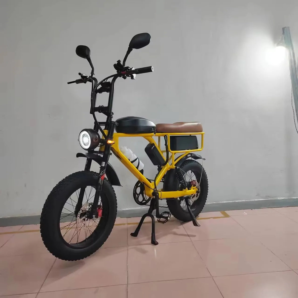 A yellow and black electric fat-tire bicycle with a brown saddle, photographed indoors against a plain light gray wall on a tiled floor.