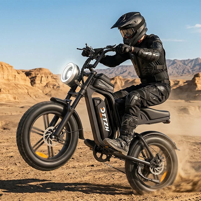 A person in full black riding gear operates a black electric motorcycle with fat tires in a sandy desert landscape under a blue sky. The bike has a round headlight, a sleek dark body, and the brand name 'HELIX' printed vertically on the side panel.