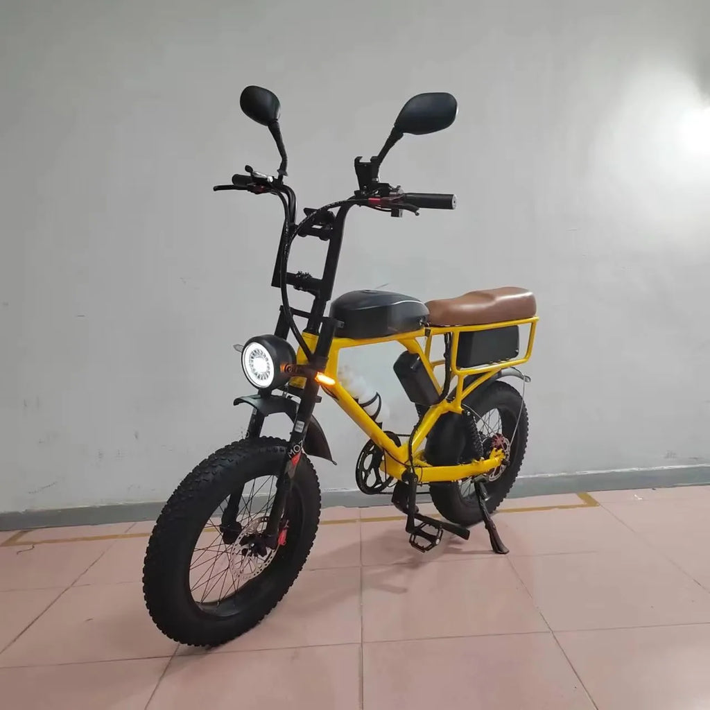A yellow and black electric fat-tire bike with a brown saddle, black mirrors, and a headlight, standing on its kickstand in an indoor setting with a tiled floor and plain wall.