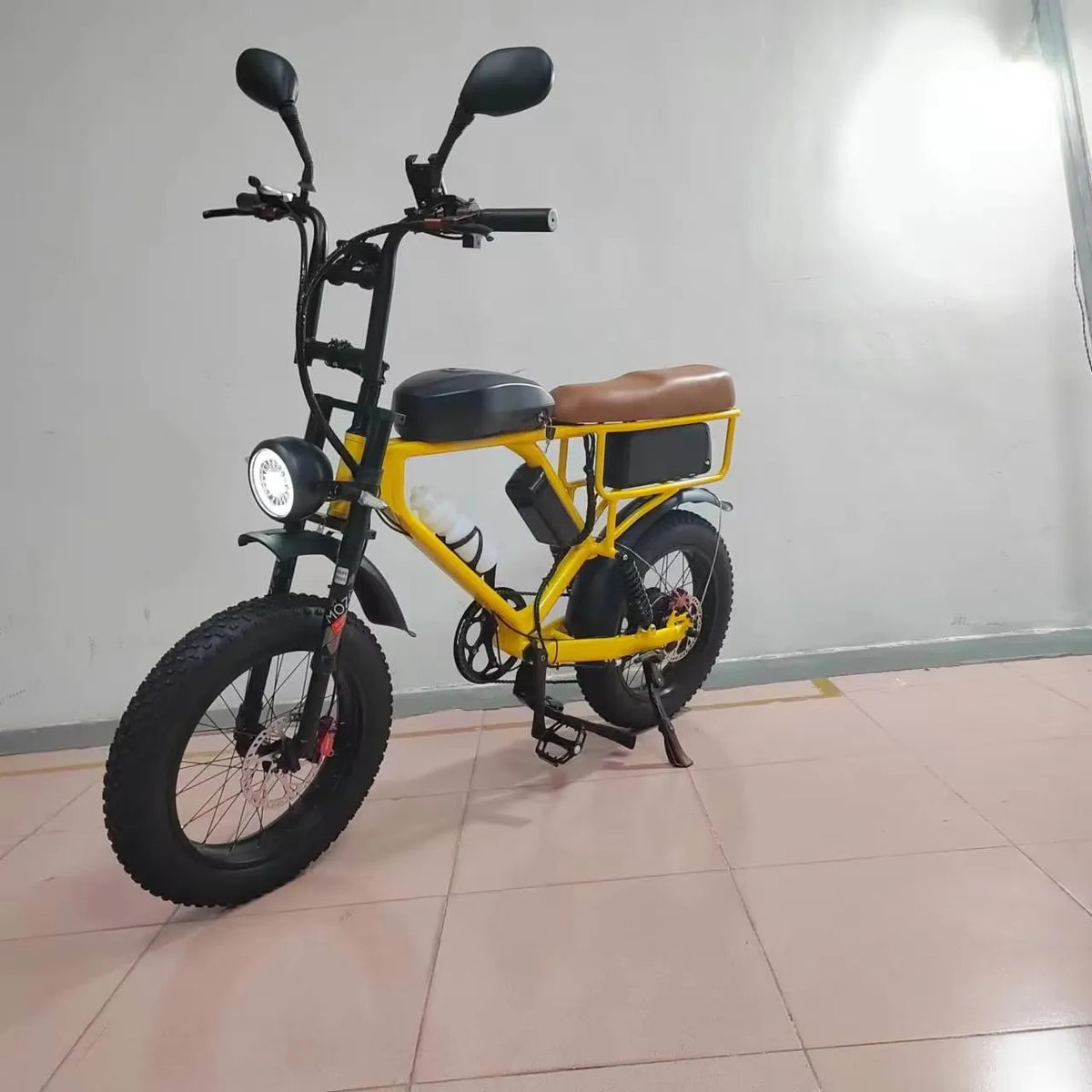 A yellow and black mini electric bicycle with fat tires, a brown saddle, and a rear cargo rack, parked indoors on a tiled floor against a plain wall.