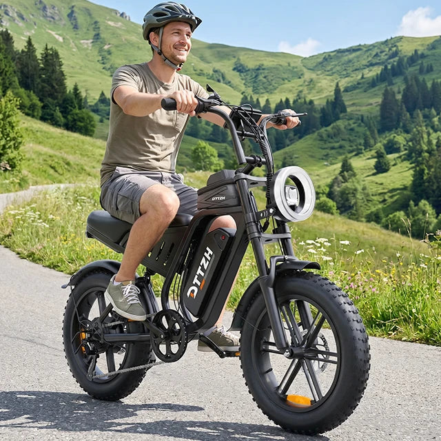 A man wearing a black helmet rides an electric bicycle on a paved mountain road. The bike is black with a round headlight, thick tires, and the brand name 'HETC' visible on the frame.