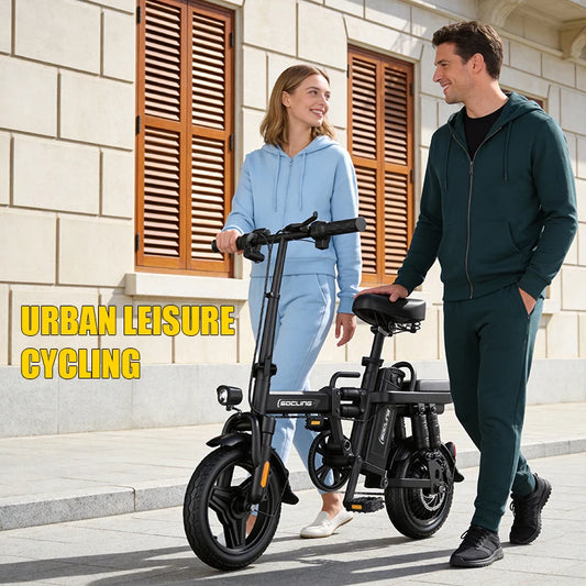 A man and a woman stand on a city sidewalk beside a compact black electric bicycle, smiling and talking. The woman wears a light blue hoodie and sweatpants, and the man wears a dark green zip-up hoodie and matching pants.