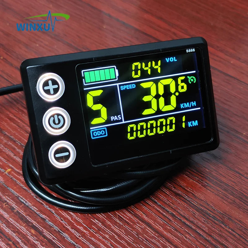 A black digital motorcycle speedometer with a backlit LCD screen showing green and yellow digits, featuring three white control buttons on the left, resting on a dark wood surface.