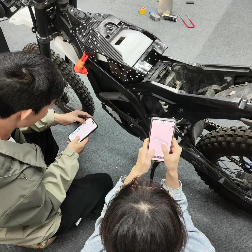 Two people holding smartphones and looking at a partially assembled black electric motorcycle in a workshop setting.