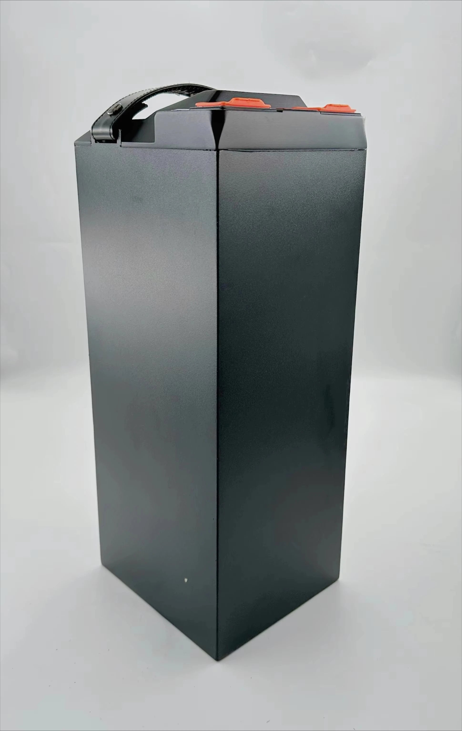 A tall rectangular black battery with a top handle and small orange terminal caps, photographed on a plain light gray studio background.