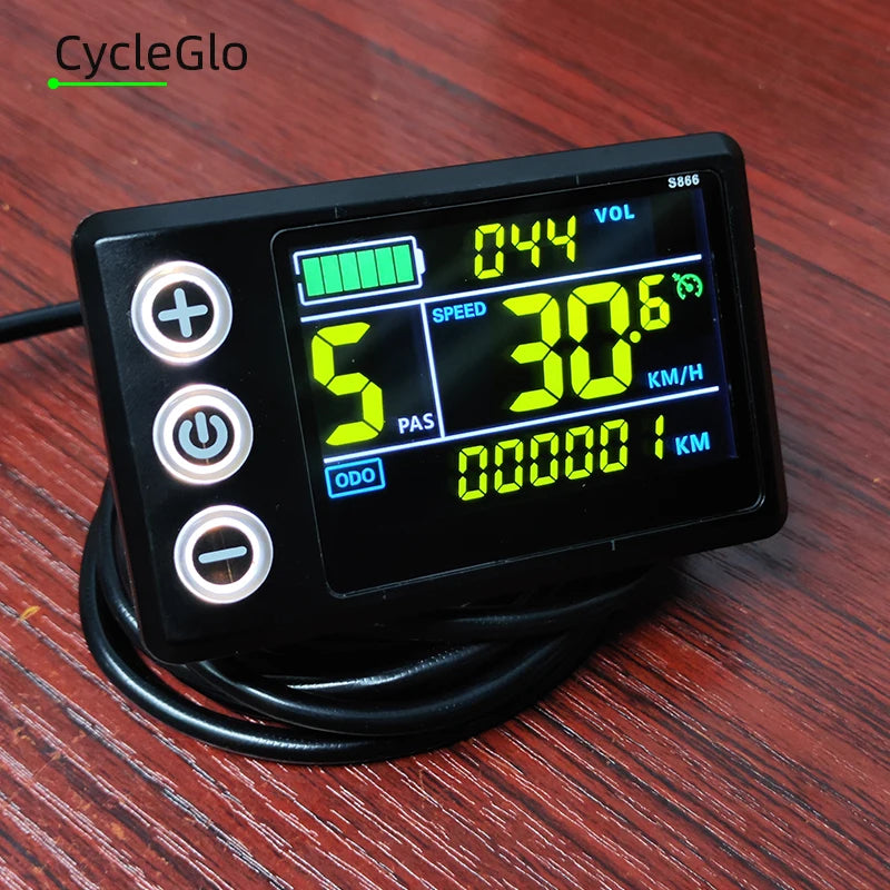 A black digital motorcycle speedometer with a backlit LCD screen showing green and yellow digits, featuring three white control buttons on the left, resting on a dark wood surface.